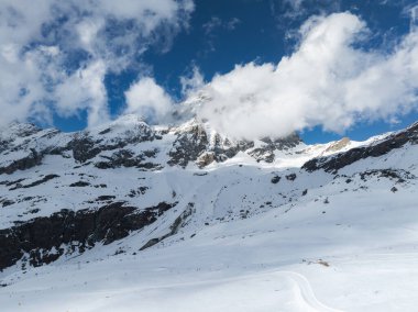Matterhorn, kar tarlaları ve engebeli kayalarla çevrili bulutların arasından yükseliyor. Kayakçılar parlak mavi bir gökyüzünün altında karla kaplı yamaçları aşıyorlar..