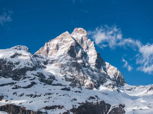 Alplerdeki ikonik Matterhorn dağında karlı bir piramit şekli, engebeli yamaçlar ve dağınık bulutlu açık mavi bir gökyüzü bulunur..