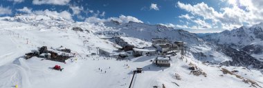 Kayak asansörleri, kayakçılar, binalar ve kırmızı kar temizleme aracı olan karlı bir kayak merkezi. Mavi gökyüzünün altındaki engebeli tepeler ve Matterhorn görülebilir..
