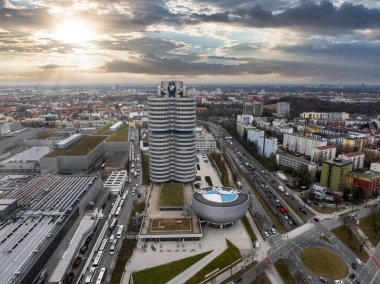 Almanya 'nın Münih kentsel manzarası, gün batımında BMW Kulesi, BMW Müzesi, kentsel binalar, yollar ve gökyüzünde dramatik güneşli bulutlar.