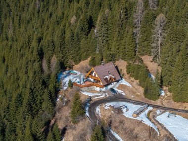 Kalın çatılı ve geniş pencereli, sık yeşilliklerle çevrili lüks bir dağ evi tarzı otel. Karlı bölgeler ve dolambaçlı bir yol sahneyi tamamlıyor.