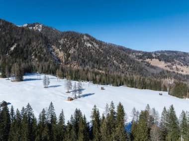 Dağılmış ahşap kulübelerle kaplı karlı bir arazi, sık yeşil ormanlarla çevrili ve Braies Gölü yakınlarında açık mavi bir gökyüzünün altında engebeli dağ yamaçları var..