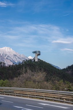 Avusturya 'nın Innsbruck kentindeki Bergisel Kayak Atlayışı, arka planda Alpler olan bir orman tepesinde duruyor. Ön planda bariyerli bir yol görülebilir..