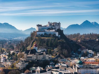 Avusturya 'nın Salzburg kentinin havadan görünüşü, bir tepede Hohensalzburg Kalesi, geleneksel Avrupa mimarisi ve açık mavi gökyüzü altındaki Alpler.