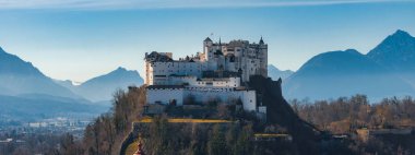 Salzburg, Avusturya 'daki bir tepenin tepesinde beyaz duvarları, kuleleri, kırmızı ve beyaz bir bayrağı ve arka planda karla kaplı dağları olan Hohensalzburg Kalesi..