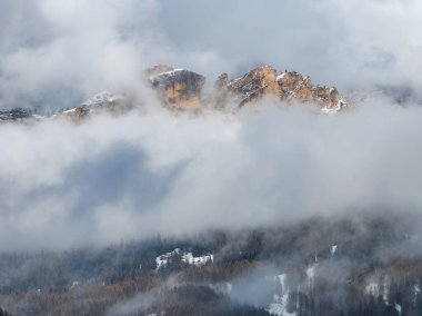 Dolomitlerin sarp kayalık zirveleri kalın bulutların arasından yükseliyor, kar tozlu zirveler ve aşağıdaki ormanlık alanlar dramatik manzaraya derinlik katıyor..