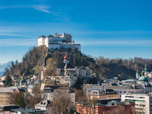 Avusturya 'nın Salzburg kentinin hava manzarasında Hohensalzburg Kalesi, kırmızı bir kilise, geleneksel binalar ve kar kaplı dağlar yer alıyor..