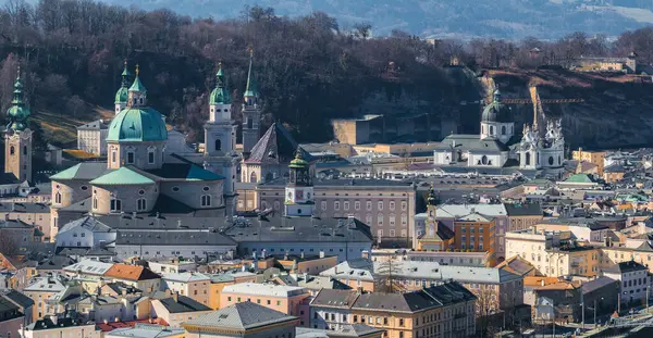 Salzburg, Avusturya 'nın havadan görünüşü, yeşil kubbeli Salzburg Katedrali, pastel renkli tarihi binalar ve arka planda ağaçlık bir tepe.