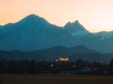 Neuschwanstein Şatosu gün batımında, karla kaplı Bavyera Alpleri ile çevrili olarak sıcak bir şekilde parlar. Kalenin altında dağınık çatıları olan bir köy var..