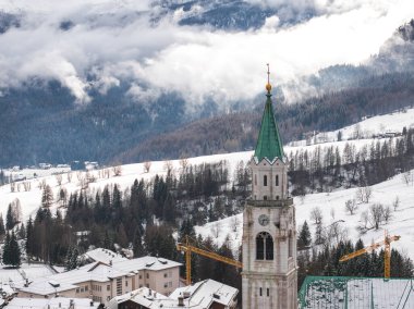 İtalya 'nın Cortina dAmpezzo kentinin havadan görünüşü, bazilika Minore çan kulesi, karla kaplı çatılar, turnalar ve bulutlu Dolomite dağları.