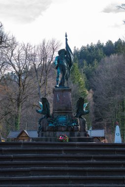 Üzerinde ejderha motifleri olan taştan bir heykel taşıyan bronz asker heykeli ağaçlar, taş basamaklar ve Avusturya 'nın Innsbruck kentindeki üssünde bir çelenk ile çevrilidir.
