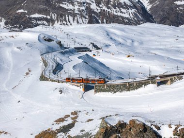 Bir turuncu tren, arka planda engebeli zirveleri olan İsviçre 'nin Zermatt kentindeki Gornergrat Demiryolu' nun eğri demiryolu köprüsünde seyahat eder..