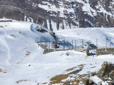 İsviçre, Zermatt yakınlarındaki karlı alp manzarasında Gornergrat Tren İstasyonu, raylardaki kırmızı tren, engebeli dağlar ve dağınık kayakçılar yer alıyor..