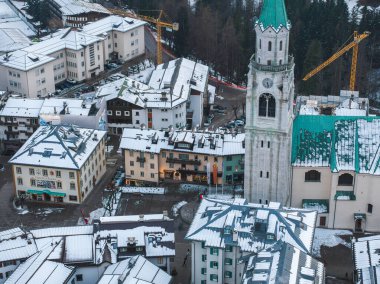 Dolomitlerdeki Cortina ve Ampezzo 'nun havadan görünüşü, Bazilika Minore çan kulesi, karla kaplı çatılar ve arka planda inşaat vinçleri yer alıyor..