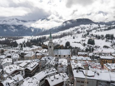 İtalya 'nın Cortina dAmpezzo kentinin hava manzarasında yeşil kulesi, dağlık binaları, karlı yamaçları ve 2026 Kış Olimpiyatları için inşaatı bulunan bir kilise yer alıyor..