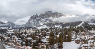İtalya 'daki Cortina dAmpezzo' nun havadan görünüşü, karla kaplı çatılar, dağlık binalar, yemyeşil ağaçlar ve Dolomitler kısmen bulutlarda zirve yapıyor..