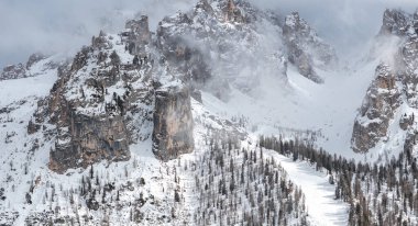 İtalya 'nın Cortina kentindeki Dolomitlerin tepelerini kaplayan kar, sisli, kayalık uçurumlar ve ön planda seyrek kar tozlu ağaçlarla kaplı bir kayak yamacı..