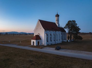 St. Coloman Kilisesi açık arazileri, uzak tepeleri ve yumuşak bir akşam gökyüzü olan kırsal bir arazide duruyor. Yolun yakınına bir araba park etmiş..