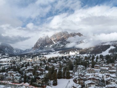 Karla kaplı çatılar, yemyeşil ağaçlar ve Dolomite tepeleri Cortina ve Ampezzo 'nun etrafını sarar. 2026 Kış Olimpiyatları için kayak pistleri ve inşaat görünür.