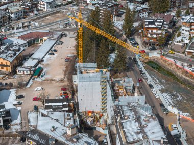 Sarı bir vinç İtalya 'nın Cortina kentindeki bir inşaat sahasının üzerinde duruyor. Etrafı karla kaplı çatılar, dağlık binalar, nehir ve dağ manzaralarıyla çevrili..