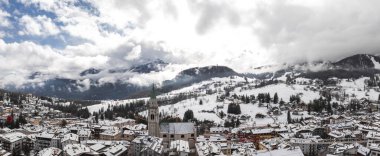 İtalya 'nın Cortina dAmpezzo kentinin havadan görünüşü, karla kaplı çatılar, yeşil spiral bir kilise, dağ zeminleri ve 2026 Kış Olimpiyatları için yapılan inşaatı gösteriyor..