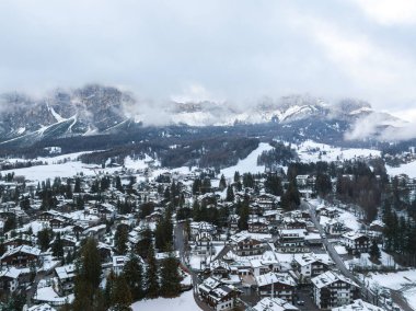 İtalya 'daki Cortina dAmpezzo' nun havadan görünüşü, karla kaplı alp mimarisi, sık ormanlar, kayak pistleri ve bulutlu gökyüzünün altında Dolomitler yer alıyor..