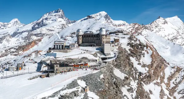 İsviçre 'nin Zermatt kentindeki Gornergrat Gözlemevi' nin hava manzarası. Karla kaplı tepeler, Matterhorn, bir tren istasyonu ve parlak turuncu bir tren istasyonu..