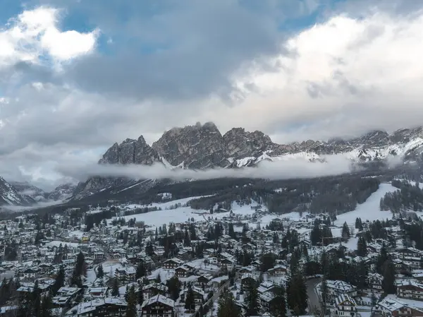 İtalya 'nın Cortina dAmpezzo kentinin karla kaplı çatıları, dağlık binaları, dolambaçlı yolları ve Dolomitlerin kısmen bulutlarla örtülü olduğu hava manzarası..