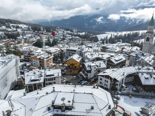 İtalya, Cortina dAmpezzo 'nun hava görüntüsü, karla kaplı çatılar, sarı bir bina, bir kilise çan kulesi, bir vinç ve etrafındaki dağları gösteriyor..