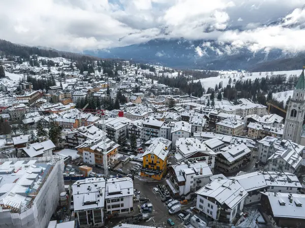 İtalya 'nın Cortina dAmpezzo kentinin havadan görünüşü, arka planda kar tozlu çatılar, sarı bir bina, bir kilise kulesi ve Dolomite dağlarını gösteriyor..