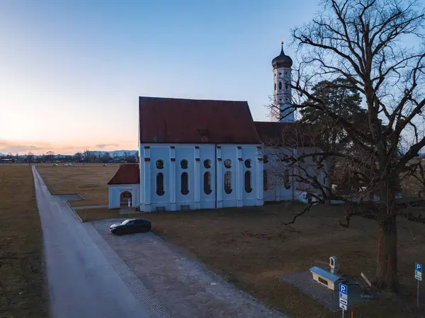 Kırmızı çatılı beyaz bir kilise ve süslü çan kulesi açık alanlarda duruyor. Çevrede asfalt bir yol, park edilmiş bir araba ve büyük yapraksız bir ağaç var..