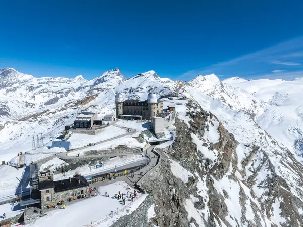 Zermatt yakınlarındaki karlı bir sırt üzerindeki Gornergrat Gözlemevi 'nin havadan görünüşü. Matterhorn ve pürüzsüz alp zirveleri berrak mavi gökyüzünün altında..