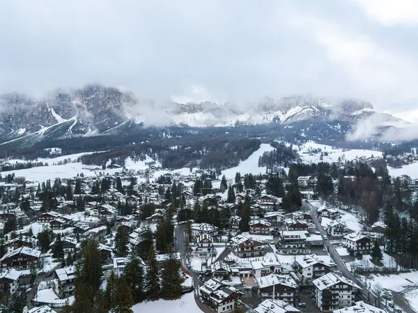 İtalya 'daki Cortina dAmpezzo' nun havadan görünüşü, karla kaplı alp mimarisi, sık ormanlar, kayak pistleri ve bulutlu gökyüzünün altında Dolomitler yer alıyor..