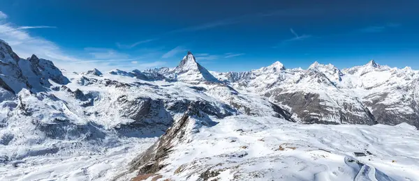 Mavi gökyüzünün altında engebeli dağlarla çevrili Matterhorn 'un hava manzarası. Kayak izleri ve gözlemevi olan karlı bir manzara görülebilir..