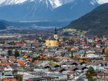 Sarı kilisesi ve yeşil çan kulesi olan bir Avusturya kasabası. Kırmızı çatılı binalar, yemyeşil tepeler ve karlı dağlarla çevrili..