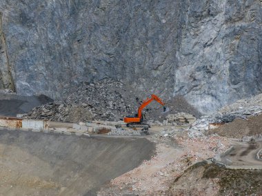 Turuncu bir kazıcı Avusturya Alpleri 'ndeki kayalık bir inşaat sahasında çalışıyor. Etrafı moloz, gevşek kayalar ve arka planda dik bir yamaç ile çevrili..