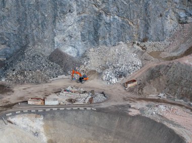 Avusturya Alpleri 'ndeki bir inşaat alanında turuncu bir kazıcı çalışıyor. Etrafı moloz, toprak yollar ve engebeli arazideki sarp kayalıklarla çevrili..