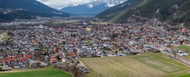 Kırmızı çatısı ve modern binaları olan bir Avusturya kasabası. Kubbesi olan merkezi bir kilise. Tepe üstü bir şato ve arka planda kar kaplı Alpler..