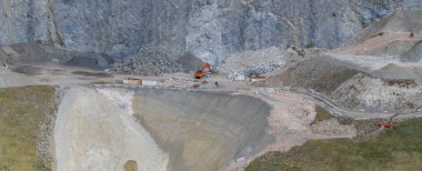 Avusturya Alplerinde, dik yamaçlar, toprak yollar ve dağınık malzemelerle çevrili kayalık bir inşaat sahasında turuncu bir kazıcı çalışıyor..