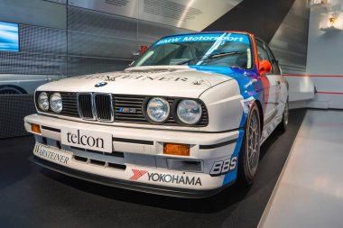 Beyaz gövdeli bir BMW M3 E30 yarış arabası, BMW Motorsport üniforması ve Münih 'teki BMW Müzesi' nin modern iç mekanında sergilenen sponsor logoları..