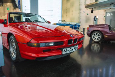 Üstü açık, parlak çizgileri ve belirgin ızgarası olan kırmızı bir BMW, klasik arabalar ve modern bir iç mekan ile çevrili BMW Müzesi 'nde sergileniyor..
