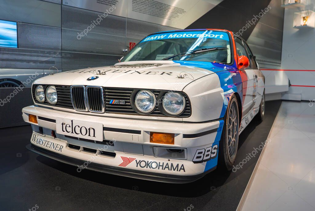 A BMW M3 E30 race car with white body, BMW Motorsport livery, and sponsor logos displayed in the modern interior of the BMW Museum in Munich.