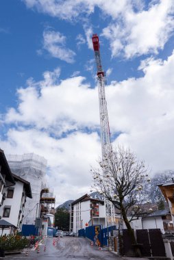 İtalya, Cortina dAmpezzo 'da bir inşaat sahasında kırmızı ve beyaz bir vinç. İskele, bariyerler, seyrek ağaçlar ve arka planda karla kaplı dağlar..