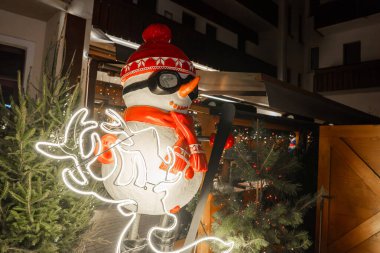 Kırmızı şapkalı, eşarplı, kayak gözlüklü neon rengeyiği ışıklarıyla çevrili ve Cortina ve Ampezzo 'daki alp tahta yapılara karşı dizilmiş yeşil ağaçlarla aydınlatılmış bir kardan adam..