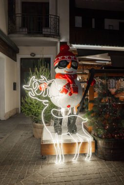Kayak, kırmızı şapka ve eşarplı bir kardan adam Cortina dAmpezzo, İtalya 'da neon bir ren geyiğinin yanında duruyor. Çimenlik ve ılık ışıklar alp büyüsü ekler..