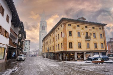 İtalya 'nın Cortina dAmpezzo şehrinde, bazilika Minores çan kulesi, armalı sarı bir bina ve Alp tarzı mimarinin yer aldığı karlı bir cadde..