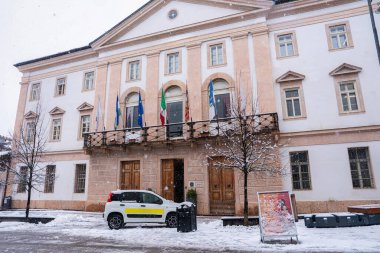 İtalya, Cortina dAmpezzo 'da balkonunda bayraklar, karla kaplı zemin, kemerli pencereler ve yakınlarda park etmiş bir araç olan neoklasik bir bina..