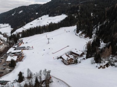 Kayak asansörlü karlı alp manzarası, yamaçlarda bir kulübe, yemyeşil ormanlar, yuvarlanan tepeler, ve Dolomitler 'de bulutlu bir gökyüzü, İtalya.