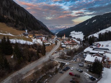 İtalya 'nın Dolomitler kentindeki bir Alp köyünün hava manzarasında yüksek kulesi, karla kaplı yamaçları, yemyeşil ormanları ve günbatımı gökyüzü olan bir kilise var..