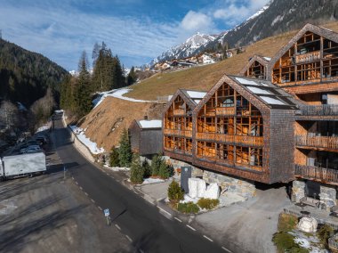 Üç köşeli çatısı ve ahşap cephesi olan Alp oteli, Dolomitler 'de bir dağ yolu boyunca kurulmuş, etrafı ağaçlar, yamaçlar ve karlı tepelerle çevrili..